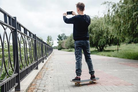 Ein Junge steht auf seinem Skateboard und macht mit seinem Handy ein Selfie.